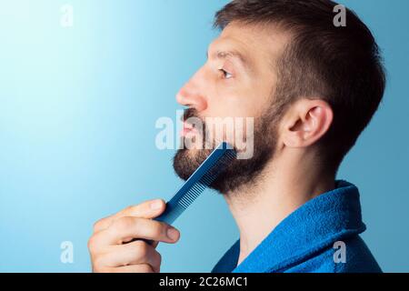Der Mann mittleren Alters kämmt seinen Bart auf blauem Hintergrund,`s Pflegekonzept des Mannes. Stockfoto